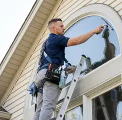 Professional window cleaner on a ladder using a squeegee to clean an exterior arched window, Scottsdale, AZ.